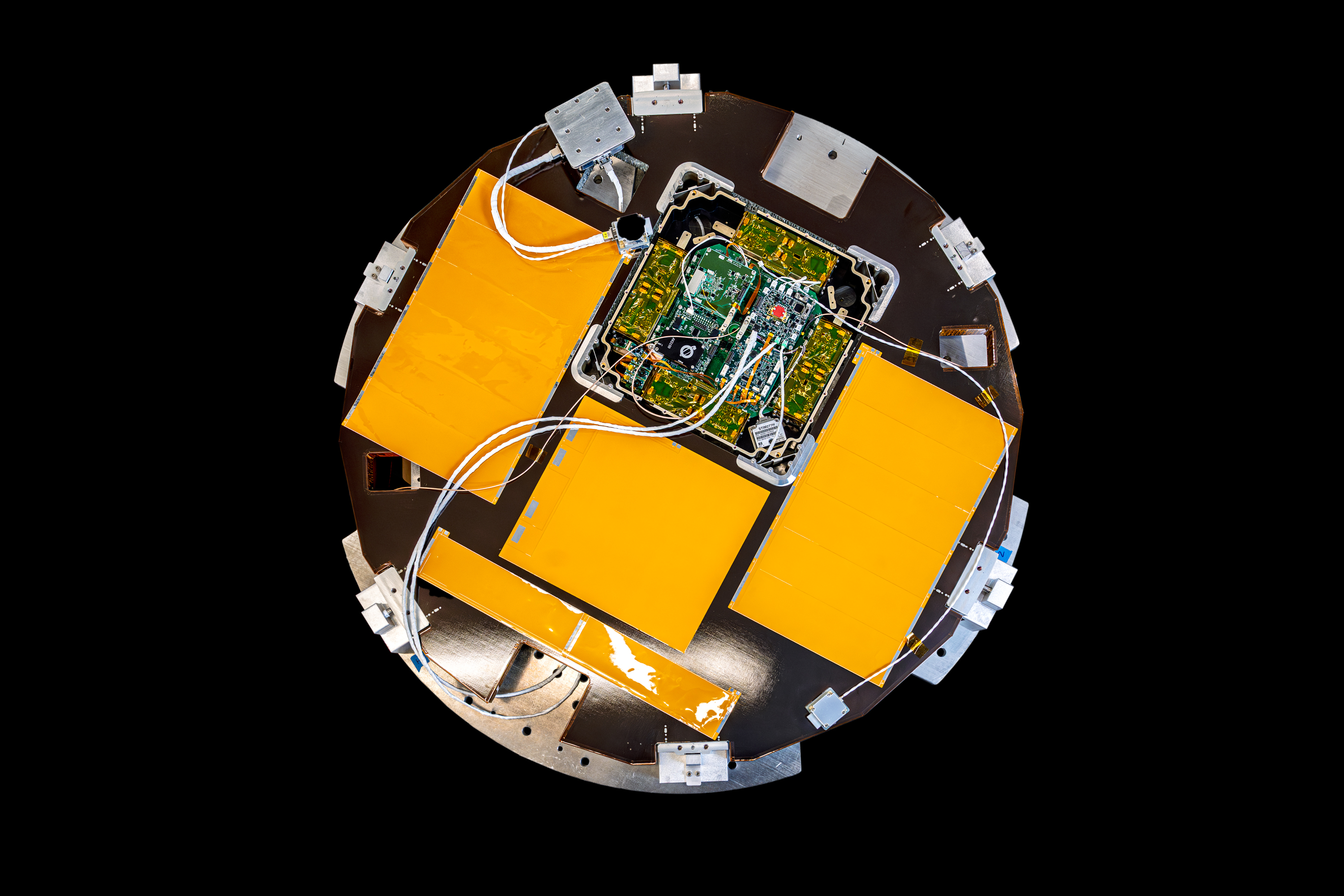 The image shows a new satellite shaped like a disk. The satellite's surface is primarily a dark color with several bright orange rectangular panels arranged symmetrically around the center. In the middle, there is a complex arrangement of electronic components and circuit boards, connected by various white cables and wires. The satellite also features several metallic attachment points and structural elements around its perimeter. The background is completely black, highlighting the satellite's design.