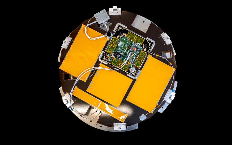 The image shows a new satellite shaped like a disk. The satellite's surface is primarily a dark color with several bright orange rectangular panels arranged symmetrically around the center. In the middle, there is a complex arrangement of electronic components and circuit boards, connected by various white cables and wires. The satellite also features several metallic attachment points and structural elements around its perimeter. The background is completely black, highlighting the satellite's design.