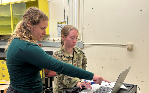 Two women, one cadet and another plain clothed pointing at a computer screen.